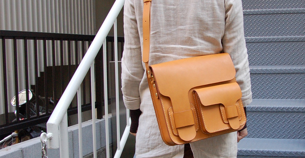 一個由棕色皮革製成的單肩包，有兩個翻蓋前袋和一個主隔層，以工整的縫線加固。