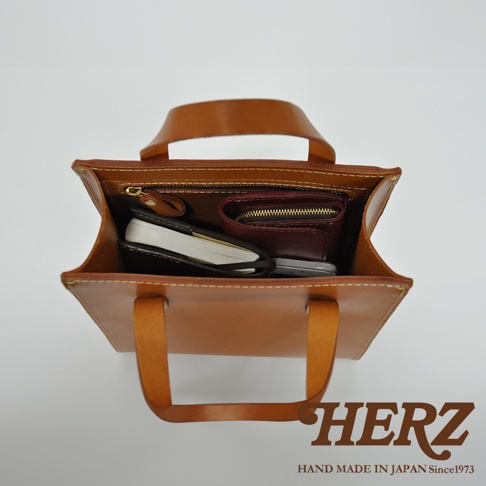 職人手作皮革直式托特包 T-44 XS，以淺棕色皮革製成，包內可見筆記本、手機及紅色零錢包，頂部有拉鍊開合及提把，整體風格簡約。