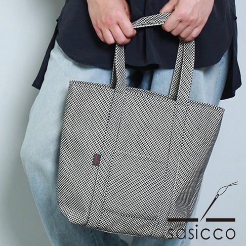 一手提著黑白格紋三河木綿刺子織OBI托特包，包包有兩個提把，正面有一個小口袋，帶有紅色標籤。