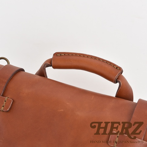 職人手作皮革水洗加工復古郵差包，特寫焦糖色皮革提把與包身細節，手工縫線清晰可見，展現高品質皮革質感。