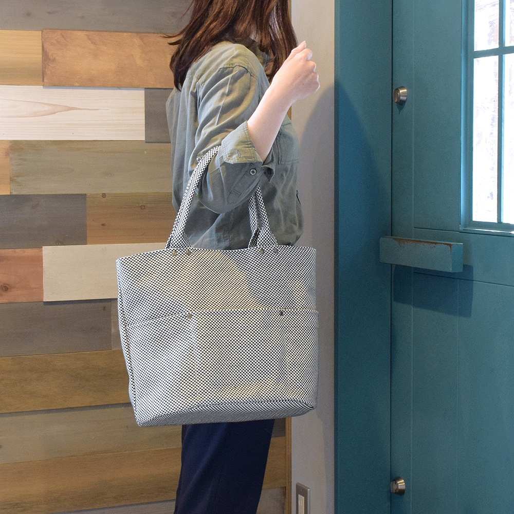 一位女性肩背一個日本製三河木綿刺子織大開口托特包，包款為黑白格紋，材質為棉，有兩個提把，包包前方有兩個貼袋。