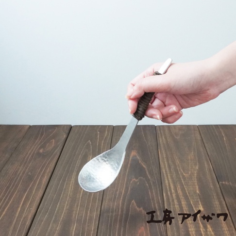 手持鋁製湯勺，勺面有錘目紋，勺柄為深棕色編織紋，置於深棕色木紋桌面。