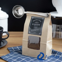 銀色不鏽鋼咖啡量匙夾，夾著印有「COFFEE MEDIUM」字樣的牛皮紙咖啡豆包。量匙夾上印有「coffee」字樣。背景有黑咖啡杯和白色咖啡濾杯。