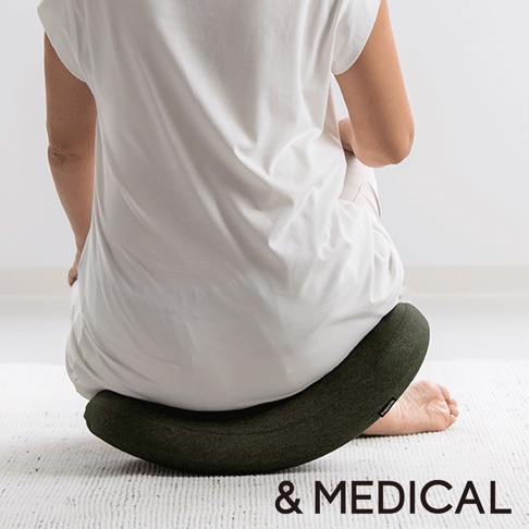 綠色U型搖搖枕，表面織紋清晰，置於白色地毯上，一位穿著白色上衣、米色褲子的人盤腿坐於其上。