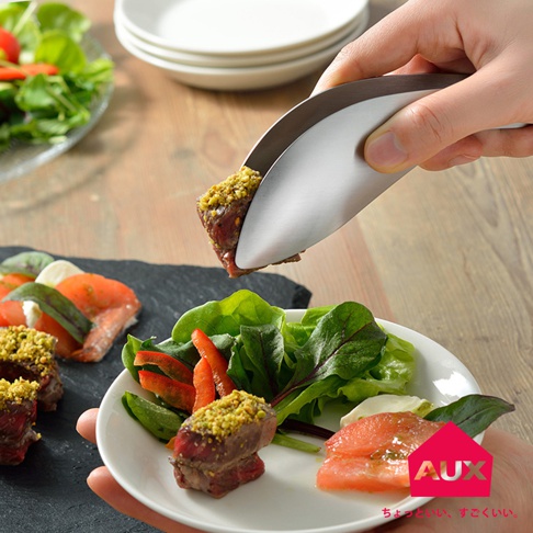 一只手持著一對銀色金屬不沾桌調理幫手夾，夾著一塊裹有綠色碎粒的牛肉。下方是裝有沙拉、番茄和牛肉的白色盤子。