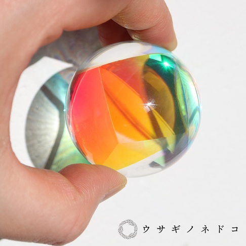 手持一顆透明的合光稜鏡球，球體表面有多個切割面，折射出紅、橘、黃、綠等多種色彩，光線透過球體產生彩虹般的效果。