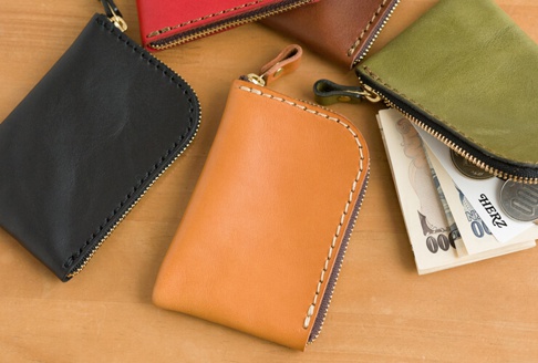 多款職人手作皮革分隔收納迷你錢包，包含黑色、橘色、紅色、棕色及綠色，皆為真皮材質，搭配金色拉鍊及手工縫線。部分錢包內有日圓鈔票與硬幣。