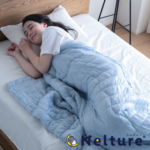 一位女子蓋著淺藍色 Nelture 涼爽夏夜舒眠單人被，被子表面有菱格紋壓線，質地輕柔，女子面露舒適睡顏。