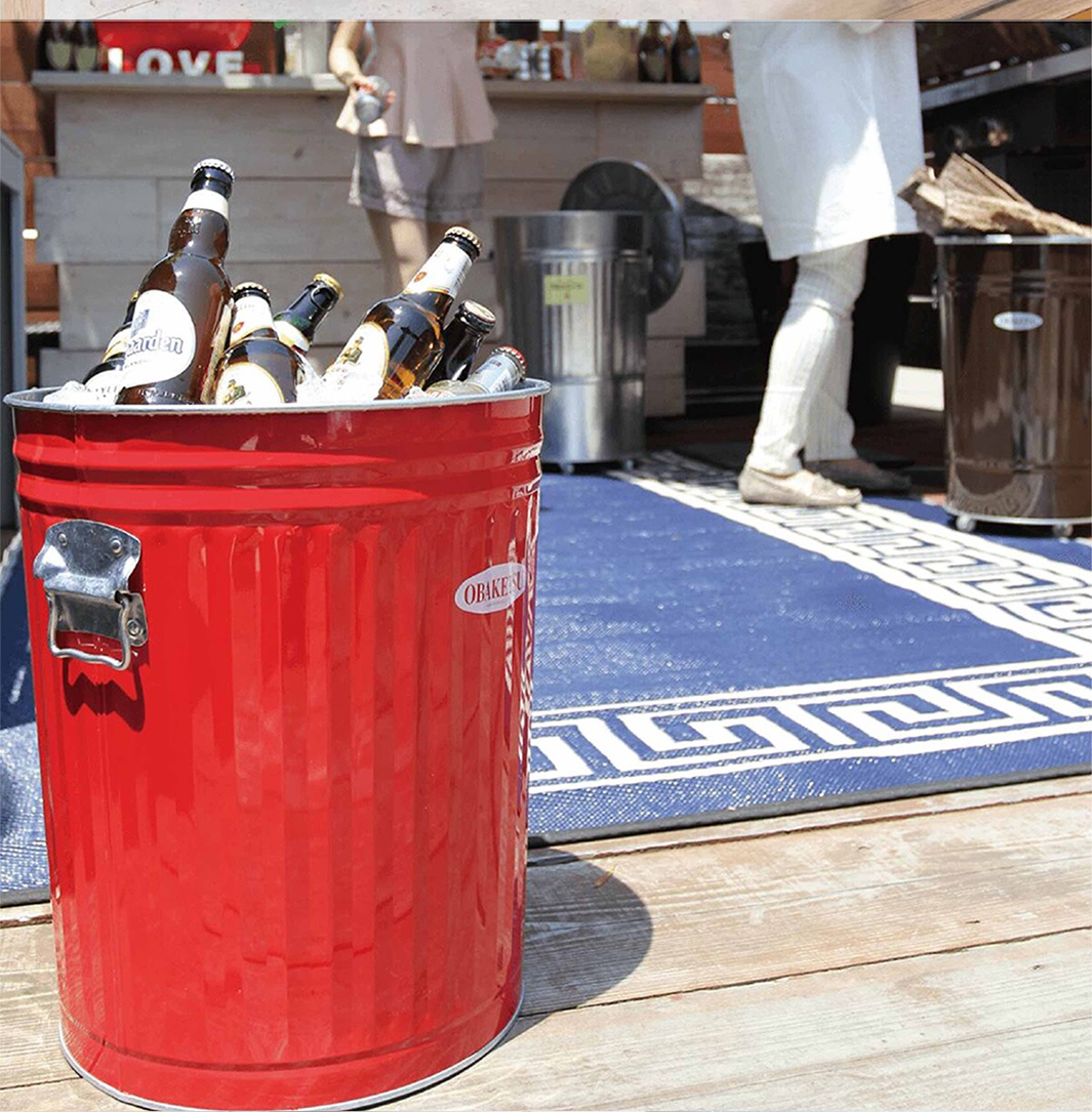 紅色 OBAKETSU 垃圾桶裝滿了冰塊和啤酒瓶，擺放在戶外的木質甲板上，背景是藍色的地毯和吧台。