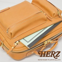 棕褐色皮革製成的職人手作兩用單肩包，採用對比色車縫線，設有拉鍊開合的前置收納袋，袋口可見部分筆記本。