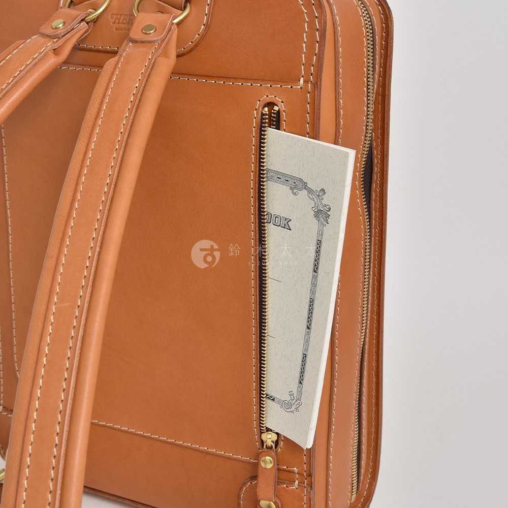 職人手作皮革商務電腦後背包 R-25，採用棕色皮革材質，車縫線清晰。背包側邊拉鍊口袋內有一本淺色筆記本。