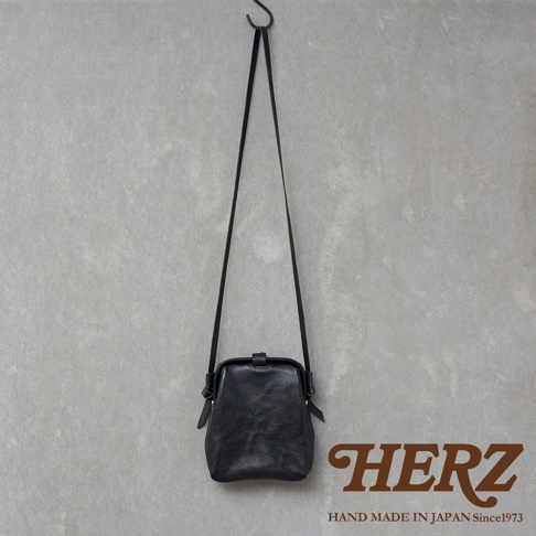 職人手作皮革Organ迷你杜勒斯側背包，黑色，荔枝紋皮革，搭配細長背帶，以金屬扣開合，懸掛於灰色紋理背景前。