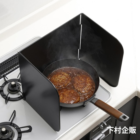 ㄇ型折疊防汙擋油板，黑色金屬材質，放置於瓦斯爐旁，圍住正在烹煮漢堡排的平底鍋，有效阻擋油煙濺出。