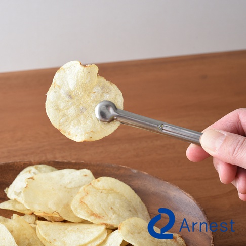 逸品物創不鏽鋼萬用料理夾，銀色，夾著一片金黃色馬鈴薯脆片，上方有木紋桌面，下方有木碗裝著多片馬鈴薯脆片。