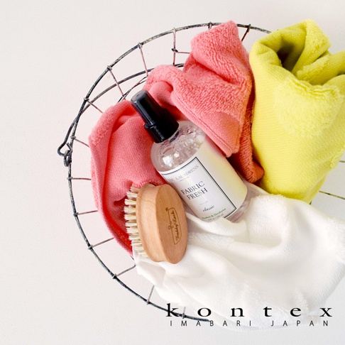 金屬籃中擺放著粉紅色、黃色和白色的毛巾，以及一瓶「THE LAUNDRESS FABRIC FRESH」噴霧和一把木柄刷。