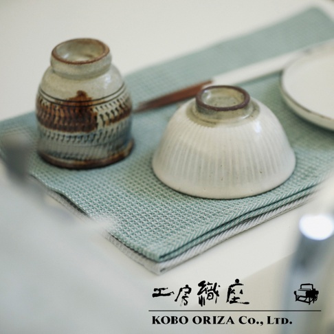 淺藍色棉麻混紡鬆餅紋拭巾，上面擺放著一個帶有棕色紋飾的陶製小罐和一個白色帶有豎紋的陶製碗。