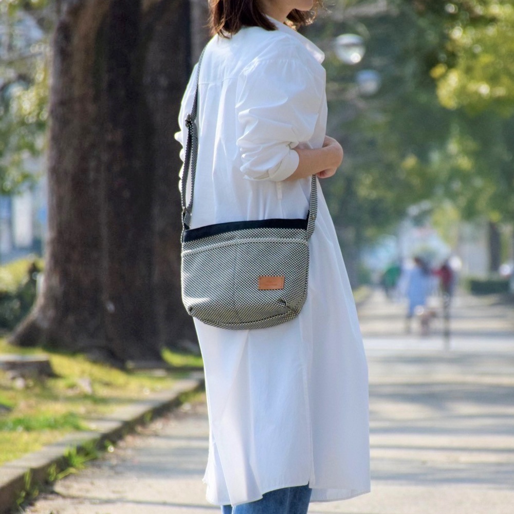 三河木綿刺子織材質的黑色與白色格紋斜肩包，配有黑色包邊及棕色皮革標籤，由一位身穿白色洋裝的女士背在肩上。