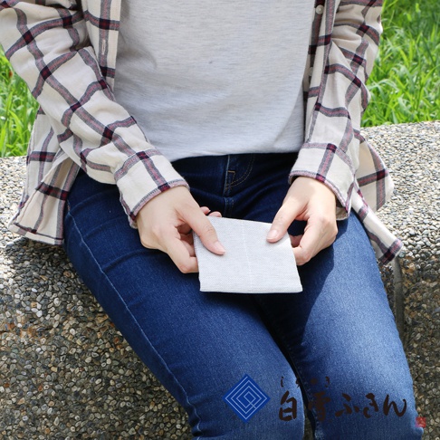 一位女士手持淺灰色方巾，方巾表面有細緻的織紋。她身穿藍色牛仔褲和一件米白色、帶有紅棕色格紋的長袖襯衫，襯衫內搭灰色T恤。