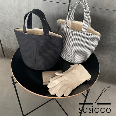 兩款三河木綿刺子織圓底水桶包，一款為黑色，另一款為黑白格紋。兩款包皆配有提把，內襯為米白色。旁邊擺放著一個米色皮革短夾及一雙米色皮革手套。