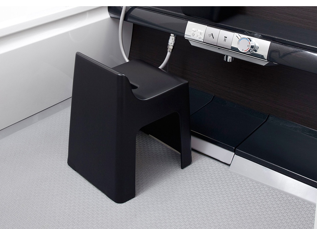 黑色一體成形簡約椅凳，以塑膠材質製成，造型為U型結構，椅面帶有輕微弧度，放置於淺灰色網格紋地板上，背景為深木紋裝飾面板與銀色金屬控制面板。