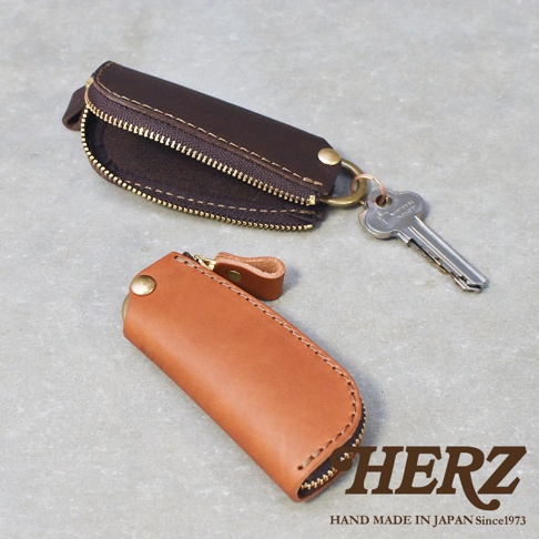 職人手作皮革Organ拉鍊鑰匙包，展示深棕色和淺棕色兩款。深棕色鑰匙包拉鍊部分打開，露出內部，並掛有一把鑰匙。淺棕色鑰匙包閉合，呈現簡潔的造型。兩款鑰匙包均採用皮革材質，配有金色拉鍊和金屬扣。