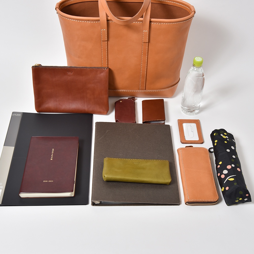 職人手作皮革經典托特包 T-9 L，配以棕色皮革化妝包、鑰匙包、卡夾、筆記本、文件夾、綠色筆袋、棕色長夾和黑色印花摺疊傘。