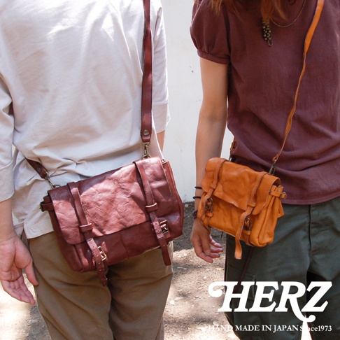 兩名模特兒展示職人手作皮革水洗加工復古郵差包，左邊模特兒背著酒紅色郵差包，右邊模特兒背著橘棕色郵差包，兩款包皆為皮革材質，帶有復古風格的設計與金屬釦環。