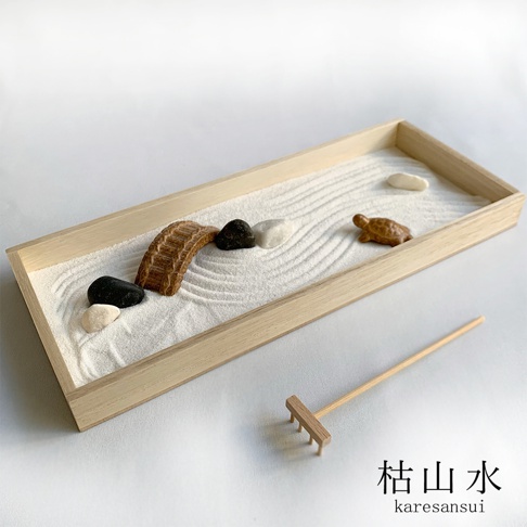 日式枯山水模型，內含白色細沙、一座木製拱橋、黑色與白色鵝卵石，以及一隻棕色小烏龜裝飾。模型置於淺色木質長方形托盤中，沙上壓有漣漪紋路，旁邊附有一支小型木製耙子。