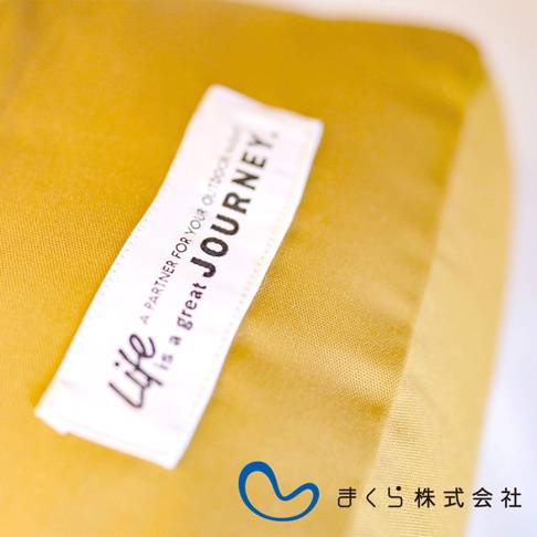 一條白色標籤縫在黃色布料上，標籤上有黑色字樣「life is a great JOURNEY」，下方有日文標誌及「まくら株式会社」。