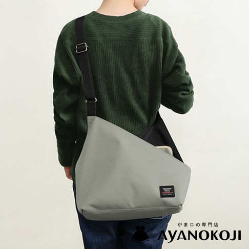 後方視角，一個人穿著綠色羅紋針織衫，肩背一個灰色方形口金單肩包，包上有一個小的黑色標籤，下方有「AYANOKOJI」的品牌標誌。