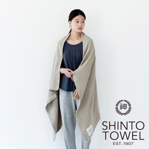 一位女性身披淺棕色2.5層紗浴巾，浴巾材質輕柔，邊緣有縫線，左下方有產品標籤。