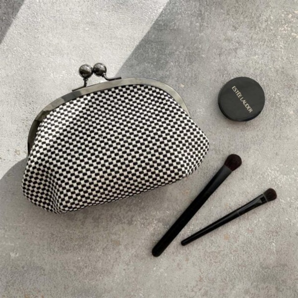 三河木綿刺子織口金零錢包，黑白格紋織紋，搭配金屬口金開合。旁邊擺放著一個黑色圓形化妝品盒及兩支黑色化妝刷。