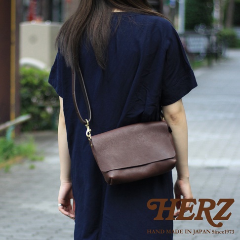 一位身穿深藍色連身裙的女性，側身展示一個精緻的棕色皮革斜背包。包包採用柔軟的紋理皮革製成，翻蓋設計簡潔大方，搭配金屬扣環增添質感。包包的背帶細長，可斜挎於肩上，展現出隨性而優雅的風格。包包右下方印有HERZ字樣及HAND MADE IN JAPAN Since1973的字樣。