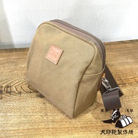 卡其色石洗帆布製成的大容量散步包，包身方正，頂部有拉鍊開合，前方有一個方形的皮革標籤，上面壓印著圖案和文字。包包側面有深棕色的帆布背帶，掛鉤為銀色金屬。背景是淺色紋理牆面和木紋桌面。右下角有兩個黑色的圖案，一個是站立的狗，另一個是兩個行李袋，下方有日文文字。