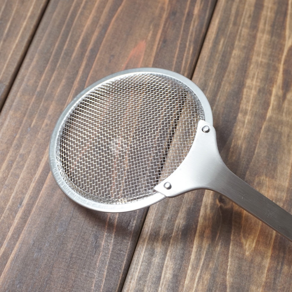 銀色不鏽鋼萬用撈油勺，帶有圓形網狀濾網和長柄，放置在深棕色木紋桌面上。
