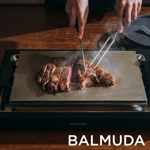 一塊切好的牛排放在淺灰色的烤盤上，烤盤置於黑色的電烤盤底座上。兩隻手持刀叉，正在切割牛排，牛排冒著熱氣。