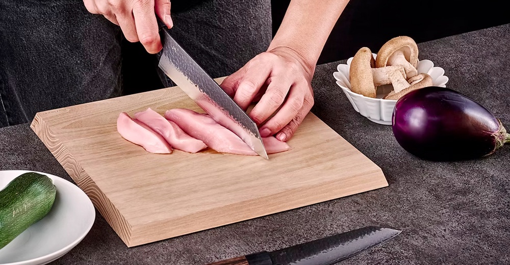 玄 / Bunka 文化刀19cm，銀色金屬刀刃，黑色木柄，正在切片粉紅色雞肉，旁邊有木質砧板、綠色櫛瓜、白色碗裡的香菇和紫色茄子。