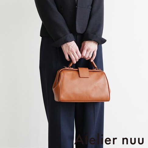 Atelier nuu 品牌標誌搭配一名身穿海軍藍西裝外套及褲裝的女子，手提一個咖啡色皮革包。