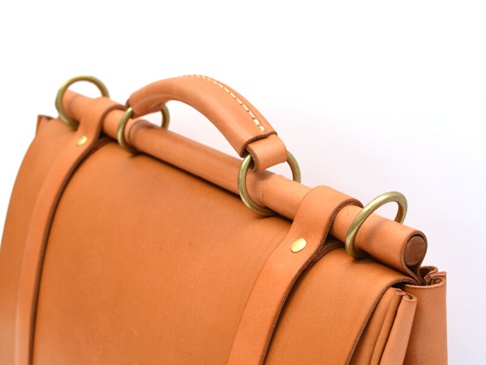 淺棕色皮革製成的三用側背包，頂部有圓形提把，以金屬環及皮革帶固定，呈現職人手作質感。