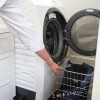 不鏽鋼製的圓形網狀收納籃，籃子裡有黑色及藍色衣物，籃子置於洗衣機旁，一名身穿白色上衣的人正伸手觸碰籃子。