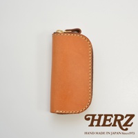 職人手作皮革橢圓形拉鍊鑰匙包，附四個掛鉤，以自然色皮革製成，搭配金色拉鍊和深棕色拉鍊頭，縫線細緻，底部有HERZ品牌標誌和HAND MADE IN JAPAN Since1973字樣。