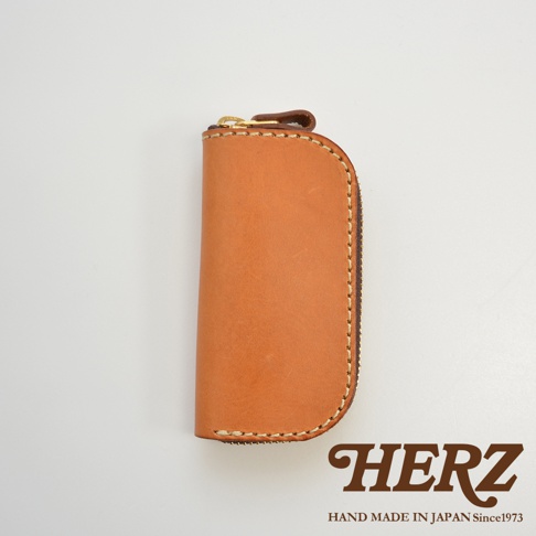 職人手作皮革橢圓形拉鍊鑰匙包，附四個掛鉤，以自然色皮革製成，搭配金色拉鍊和深棕色拉鍊頭，縫線細緻，底部有HERZ品牌標誌和HAND MADE IN JAPAN Since1973字樣。