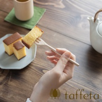 一隻手拿著一根淺色楓木點心叉，叉著一塊金黃色的蛋糕。背景有淺灰盤子裝著幾塊蛋糕，綠色杯墊上的木杯，以及白色茶壺。