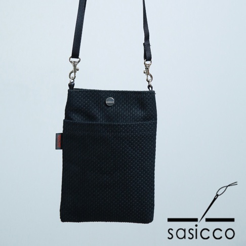 黑色三河木綿刺子織手機包，帶有可調節的黑色織帶和金屬掛鉤，正面有一個小口袋和一個帶有品牌標誌的按扣。