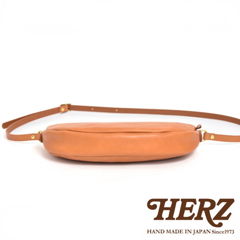 日本HERZ手工皮革半圓馬鞍包，皮色自然，配有可調節的細長皮質背帶，展示了品牌標誌和「HAND MADE IN JAPAN Since1973」的字樣。