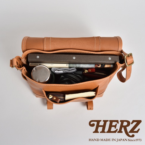 職人手作皮革復古郵差包，採用鞣製皮革製成，呈現自然焦糖色。包包內裝有書籍、保溫瓶、錢包等物品，側面有兩個小型外袋，其中一個裝有筆記本和零錢包。包包設計簡潔，適合日常攜帶。