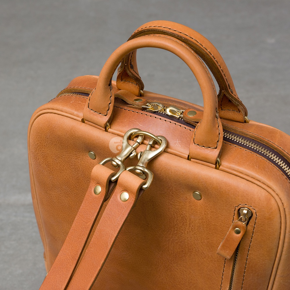 職人手作皮革Organ商務後背包，呈現溫潤的焦糖棕色，可見其細緻的皮革紋理、雙向拉鍊設計，以及可拆卸的肩背帶扣環，展現精緻工藝。