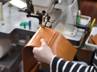 職人正在使用縫紉機製作一個棕色皮革中型雙折短夾，可見精緻的縫線與卡槽。