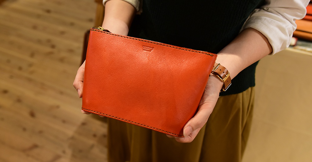 一位女士手持一個橘紅色皮革三角收納包，包包上有金色拉鍊，皮革材質，邊緣有縫線。