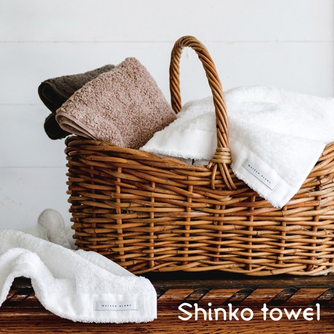 一個籐籃裡有三條毛巾，分別為棕色、米色和白色。其中白色毛巾上有 MAISON BLANC 標籤。一條白色毛巾放在籃子前面，同樣帶有 MAISON BLANC 標籤。